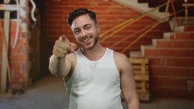 Sakallı genç adam kameraya gülümsüyor ve arka planda açık tuğla ve ekipmanla kapalı bir inşaat alanında rahatlamış bir atmosfer izlenimi veriyor..