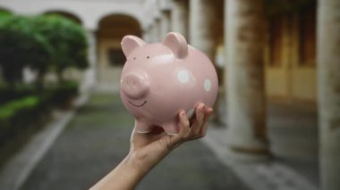 Eski bir üniversite avlusunda el ele tutuşan pembe domuz kumbarası tarihi mimarinin ortasındaki finansal temaları vurguluyor..