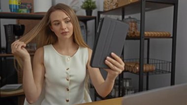 Bir ofis ortamında tablete bakarken saç fırçalayan kadın, profesyonel bir çalışma ortamında rafları ve dekorasyonu olan odak ve teknolojiyi ön plana çıkarıyor.