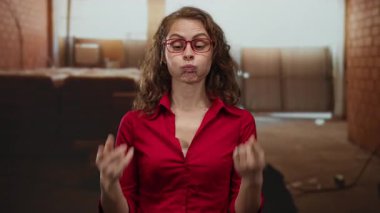 İnşaat alanında, kırmızı gömlek ve gözlük takan, bitmemiş duvarlar ve sıcak aydınlatmalarla çevrili, neşeli ve esprili bir ruh halini yansıtan komik suratlı bir kadın..