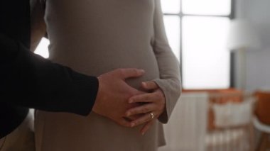 Evde eşi olan hamile bir kadın karnına dokunuyor, beklenti ve sevgi gösteriyor, sıcak odada nazik bir bağlantıyı vurguluyor..