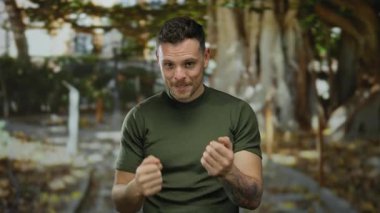 İspanyol bir adam parkta bir jest yapıyor, dövmeleri var ve yeşil bir gömlek giyiyor, doğal ortamda cazibe ve güven yayıyor..