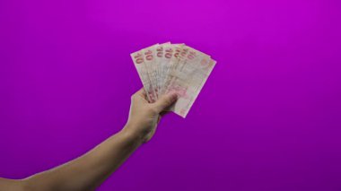 Canlı mor bir arkaplan karşısında Türk lira banknotları tutan adam, minimalist izole bir ortamda finans ve ekonomiyi vurguluyor..