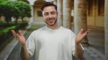 Sakallı bir adam güvenli bir şekilde şehir ortamında gülümsüyor, tarihi mimariyle çevrili, beyaz gömlek giyiyor, rahat ve enerjik tavırlar sergiliyor..