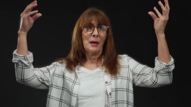 Gözlüklü son sınıf öğrencisi bir kadın, siyah bir arka plana tutkuyla işaret ediyor, ekose gömlek giyiyor, duygularını ve iletişimini izole bir ortamda ifade ediyor..