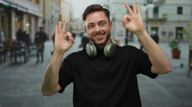 Sakallı ve kulaklıklı genç adam şehrin arka planında bulanık bir jest yaparken gülümsüyor. Açık havada tasasız ve rahat bir atmosfer izlenimi veriyor..