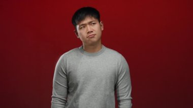 Gri süveter giymiş genç Çinli bir adam izole edilmiş kırmızı bir arka plan karşısında gülümsüyor. Mutluluk ve basitliği ifade ediyor..