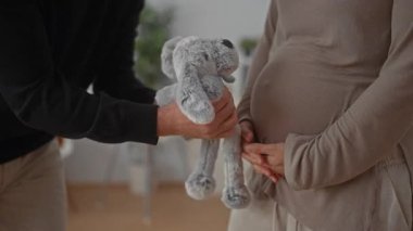 Hamile çift bir bebek odasında neşeyle etkileşime girerken, bir erkek oyuncak hediye ederek bir beşiğin yanında bir kadın bekliyor..