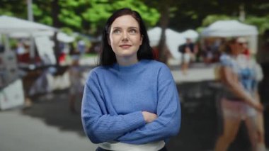 Güneşli bir günde, açık havada mavi kazaklı bir kadın arka planda market tezgahlarıyla yukarı bakıyor. Merakını dile getiriyor ve hareketli bir sokak sahnesine odaklanıyor..