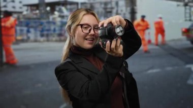 Çalışan inşaat sahasında kamerayla fotoğraf çeken kadın turuncu üniformalı ve endüstriyel geçmişli işçileri gündüz vakti yakalıyor..