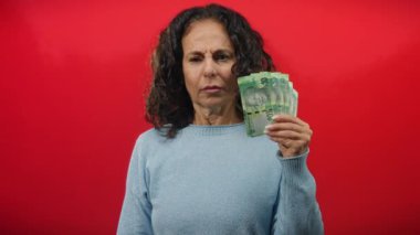 Güney Afrika banknotlarını kırmızı duvara dayayan, endişeli görünen, orta yaşlı, kıvırcık saçlı, açık mavi kazaklı bir kadın..