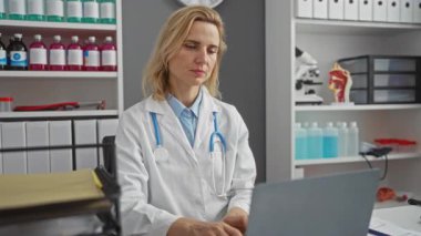 Steteskoplu beyaz önlüklü kadın, profesyonel ve odaklanmış bir çalışma ortamı sergileyen tıbbi malzemelerle çevrili bir klinikte dizüstü bilgisayar kullanıyor..