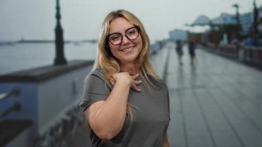 Deniz kenarındaki gezinti alanında gülümseyen sarışın genç bir kadın. Arka planı bulanık. Açık hava manzarasını gösteriyor..