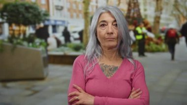 Açık bir şehir caddesinde kolları çapraz olan gri saçlı yaşlı bir kadın, şehir ortamında kendine güven ve dayanıklılık gösteriyor, pembe bir süveter ve dövme giyiyor..