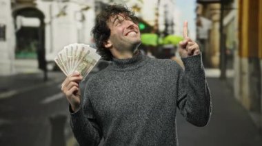 Şehir sokaklarında İspanyol bir adam elinde Rus rublesi banknotları tutuyor ve neşeli bir ifadeyle yukarı doğru işaret ediyor. Şehir ortamındaki finansal fırsatları vurguluyor..