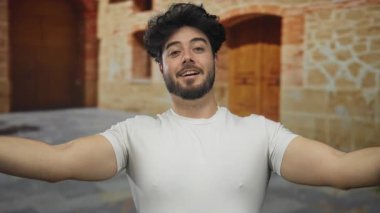 Sakallı İspanyol bir adam eski bir üniversite binasının arka planında kolları ve göğsü açık poz veriyor..