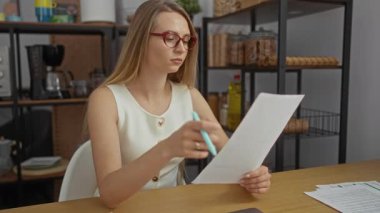 Modern ofiste masa başında kitap okuyan bir kadın gözlük takıyor ve etrafı raflar ve mutfak aletleriyle çevrili beyaz bir bluz giyiyor..