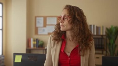 Kıvırcık saçlı, kırmızı gözlüklü bir kadın, kitap, bitki ve bilgisayar masasıyla çevrili bir ofis ortamında sakin ve kararlı bir tavır sergiliyor..