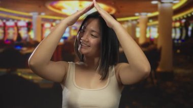 Kumarhanede genç bir kadın elleriyle çatı jesti yapıyor. Parlak ışıkların arasında gülümsüyor..