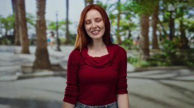 Şehir sokaklarında gülümseyen bir kadın, kırmızı giyinmiş, mutluluk ve kendine güveni gösteren, ağaçlarla çevrili, güzelliği ve çekiciliği somutlaştıran.