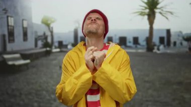 Sarı ceketli ve bereli genç adam şehrin arka planında başparmağıyla gülümsüyor açık hava coşkusu ve pozitif tavır sergiliyor..