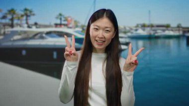 Güneşli bir günde arka planda teknelerle sahil limanında barış işareti ile gülümseyen bir kadın..
