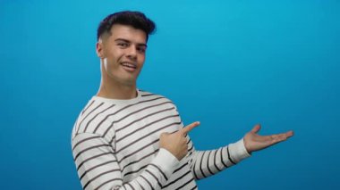 Çizgili gömlekli genç adam gülümsüyor ve mavi arka plana işaret ediyor.