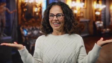 Kıvırcık saçlı İspanyol kadın otel resepsiyonunda, gözlük ve süveter takıyor, ellerini uzatıp gülümseyerek hoş geldin diyor..