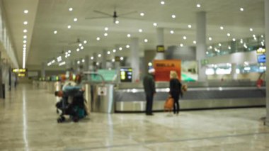 Bagajlı yolcular, parlak ışıklar ve kapalı alan ayarlarıyla bir havaalanı bagaj teslim noktasında bekliyor. Kapalı alan ayarları, odakları bozulmuş ve bulanık arka plan seyahat atmosferini yakalıyor..