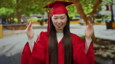 Kırmızı mezuniyet elbisesi içinde, arka planda ağaçlar ve patikalar olan bir parkta mutlu bir şekilde el kol hareketi yapan bir kadın, açık havada akademik başarı kutlamasını sergiliyor..