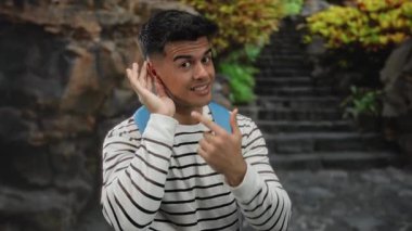 Genç adam dışarıda kulağına gülümsüyor ve el kol hareketi yapıyor. Arka planda merdivenleri olan çizgili gömlek ve sırt çantası giyiyor..