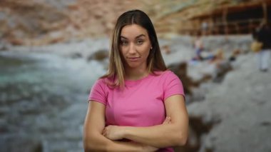 Pembe tişörtlü genç bir kadın güvenli bir şekilde sahilde duruyor, sakin bir açık hava ortamı yakalıyor, doğal güneş ışığında kayalık sahil güzelliği ile çevrili..