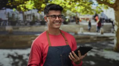 Şehrin caddesinde elinde tabletle gülümseyen genç bir adam, gözlük ve önlük takıyor. Arka planda canlı bir kalabalık ve ağaçlar var. Neşeli şehir yaşamını somutlaştırıyor..