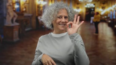 Gri saçlı bir kadın müzede el hareketi yapıyor, gülümsüyor ve sofistike bir iç mekan ortamında pozitifliği simgeliyor..