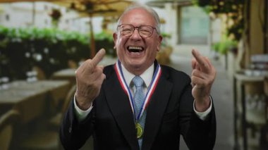 Takım elbiseli son sınıf öğrencisi, restoran terasında gülerken kaba bir jest yapıyor. Açık hava sokak ortamında, madalyayla meydan okuyan espriler sergiliyor..