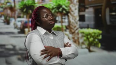 Afro-Amerikalı bir kadın canlı bir şehir caddesinde kollarını kavuşturmuş, açık şehir ortamında stil ve güç gösterisi yapıyor..