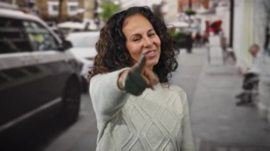 Kıvırcık saçlı bir kadın işlek bir şehir arka planında kameraya doğru gülümsüyor..