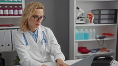 Klinikteki steteskop takan kadın doktor ciddi görünüyor, etrafında tıbbi ekipman ve dosyalarla bir masada oturuyor, profesyonel bir ortamda gözlük tutuyor..
