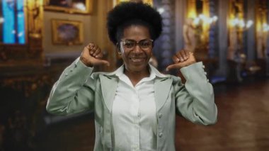 Bir müzedeki genç Afro-Amerikalı kadın kapalı bir ortamda kendini işaret ediyor, kendine güvenen ve zarif sanat eserleri ve ışıklandırmalarla çevrili..