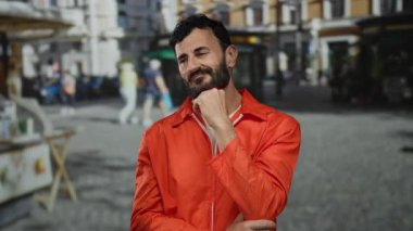 Turuncu tulumlu ve sakallı bir adam yoğun bir sokakta hapishane kıyafetleri ve kamu düzenini harmanlayarak düşünceli bir şekilde duruyor..