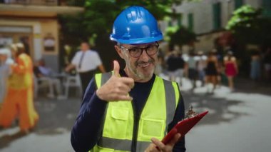 İnşaat malzemeleri giyen olgun bir adam kentin hareketli sokak ortamında güler kırmızı bir pano tutar ve bulanık bir şehir parkı zeminiyle çevrilidir..