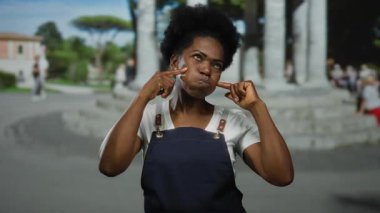 Önlüklü bir kadın açık havada gülümsüyor, bir şehir caddesinde yanaklarına dokunuyor, pozitiflik ve neşe saçıyor, şehir yaşamını ve açık hava ruhunu somutlaştırıyor..