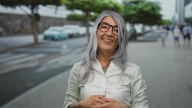 Arka planda arabalar ve ağaçlarla dolu bir caddede gülümseyen kır saçlı son sınıf öğrencisi kadın neşeli ve sıradan bir şehir manzarası sergiliyor..