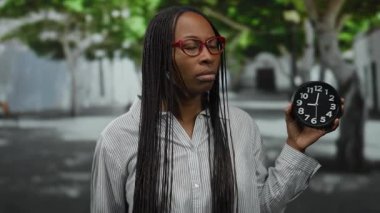 Şehir caddesinde saatli gözlüklü bir kadın, ciddi görünüyor, kıvırcık saçlı ve çizgili gömlek giyiyor, şehir ağaçlarının arasında gün ışığında duruyor..