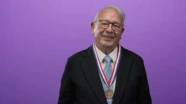 Kıdemli adam takım elbise giyiyor, neşeyle gülümsüyor, canlı mor arka plana karşı madalya sergiliyor, başarı ve memnuniyeti iletiyor..