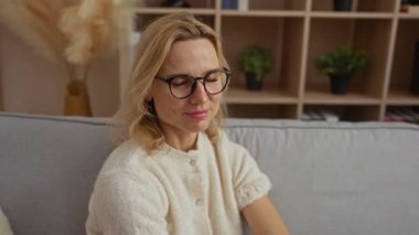 İçerde gülümseyen bir kadın rahat bir evde gözlük takıyor ve içinde bitkiler ve dekor elementleri olan bir kitaplık hazırlıyor..