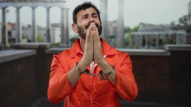 Turuncu tulumlu, sakallı ve kelepçeli İspanyol bir adam eski Roma düzeninde antik mimari arka planda el ele tutuşup yalvarıyor..