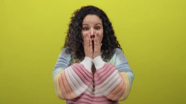 Gökkuşağı süveteri içindeki kıvırcık saçlı İspanyol kadın, canlı sarı bir arkaplan karşısında şaşkınlık gösteriyor, elleri yanaklarında duyguları iletiyor..