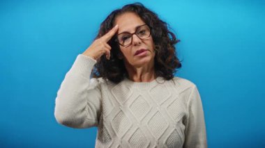 Kıvırcık saçlı, gözlüklü ve mavi arka planda düşünceli bir ifadeye sahip süveter giyen İspanyol bir kadın..