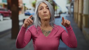 Pembe tişörtlü yaşlı kadın bulanık binalarla şehir caddesinde duruyor; sokak arkaplanı hareketli şehir ortamında açık hava ortamını öneriyor.